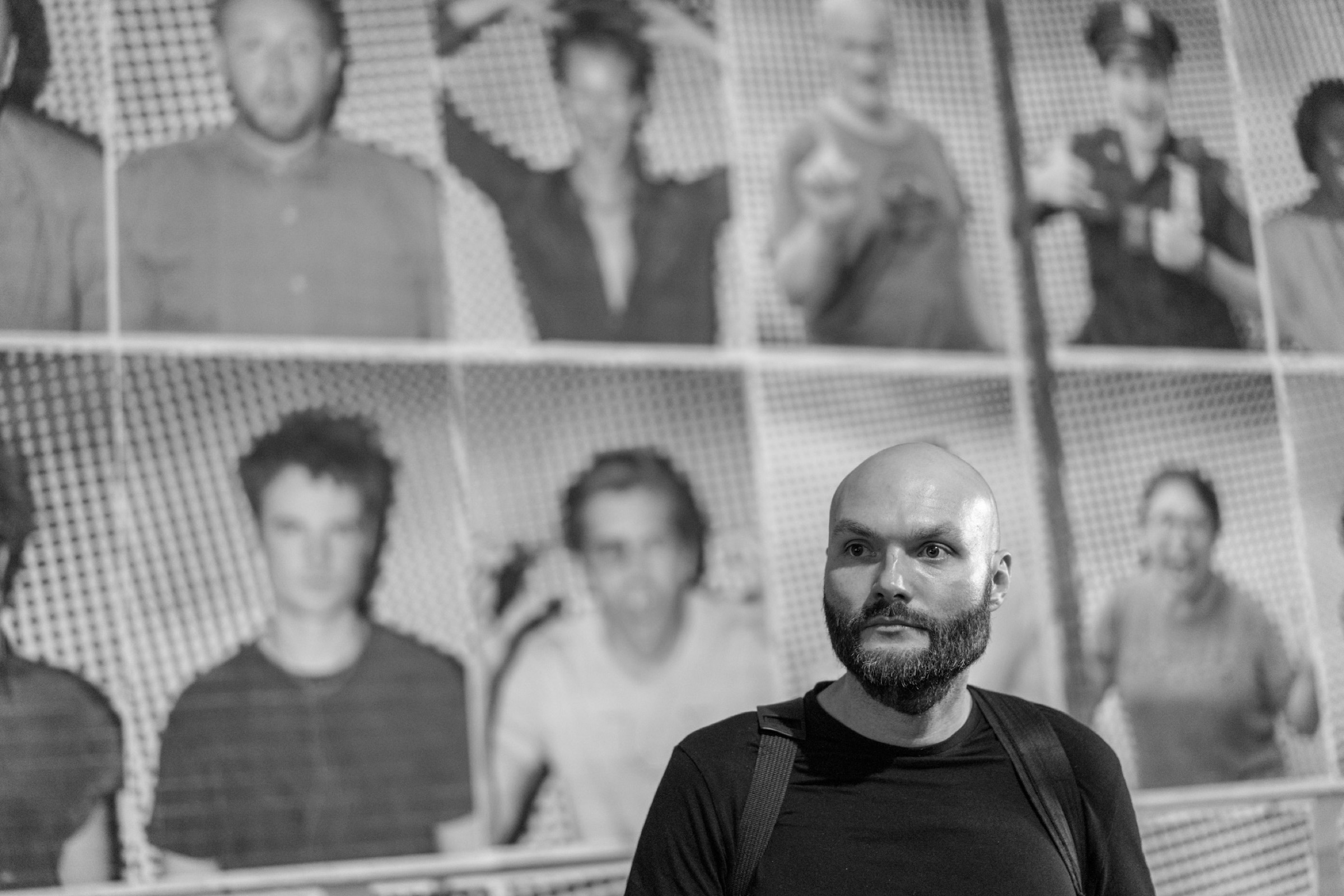 Black-and-white environmental portrait of a man standing in front of a wall of photographs